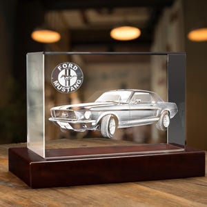 May include: A clear glass cube with a wooden base displays a 3D laser-engraved image of a classic Ford Mustang car. The Ford Mustang logo is also engraved on the cube.