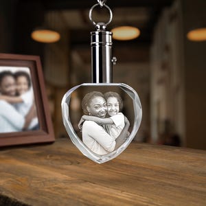 May include: A heart-shaped crystal keychain featuring a laser-etched black and white photograph of an adult and a child embracing. The crystal is attached to a polished silver-colored metal cylindrical cap with a circular ring for attachment.