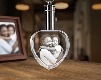 May include: A heart-shaped crystal keychain featuring a laser-etched black and white photograph of an adult and a child embracing. The crystal is attached to a polished silver-colored metal cylindrical cap with a circular ring for attachment.