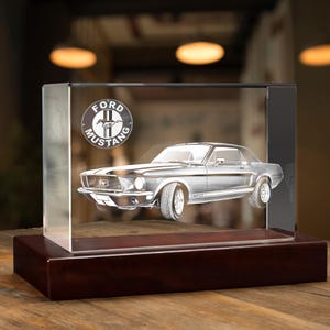 May include: A clear rectangular glass block featuring a laser-etched image of a Ford Mustang car and the circular Ford Mustang logo. The glass block is mounted on a solid, dark brown rectangular wooden base with a smooth, polished finish.