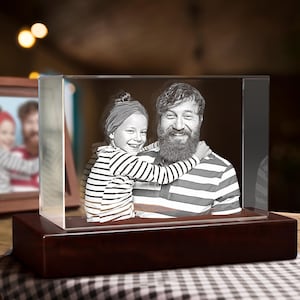 May include: A rectangular glass block featuring a laser-etched black and white portrait of a child with a headband hugging a bearded adult wearing a striped shirt. The glass block is mounted on a dark brown rectangular wooden base.
