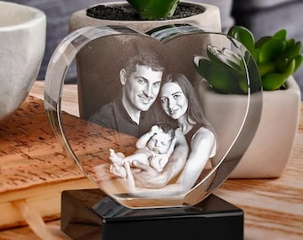 Puede incluir: Un bloque de cristal transparente en forma de corazón con una fotografía grabada con láser en blanco y negro de un hombre, una mujer y un bebé. El corazón de cristal está montado sobre una base rectangular negra sólida y decorativa.
