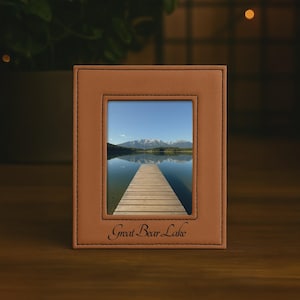 May include: A rectangular tan leather photo frame with visible stitching along the edges. The frame contains a photograph of a wooden pier extending into a calm lake toward distant mountains. The text Great Bear Lake is embossed at the bottom.