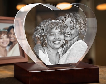 Puede incluir: Un bloque de cristal transparente en forma de corazón con una fotografía en blanco y negro grabada con láser de dos personas sonrientes con cabello rizado. El corazón de cristal está montado sobre una base rectangular de madera pulida.