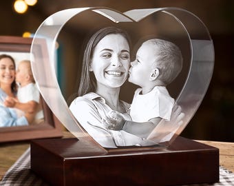 Puede incluir: Un bloque de cristal transparente en forma de corazón con una fotografía en blanco y negro grabada con láser de una persona adulta sosteniendo a un niño pequeño. El corazón de cristal se asienta sobre una base de madera oscura.