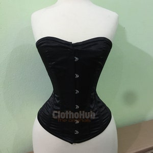 K&ouml;nnte beinhalten: Ein schwarzes, tr&auml;gerloses Korsett mit Satin-Finish und silbernen Metallverschl&uuml;ssen. Das Korsett befindet sich auf einer wei&szlig;en Schaufensterpuppe. Der Text "ClothoHub the originals" ist auf der Vorderseite des Korsetts aufgedruckt.