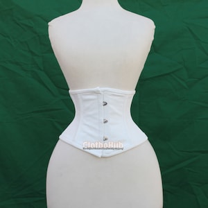Könnte beinhalten: Weißes Baumwollkorsett mit vorderem Verschluss und silbernen Knöpfen. Das Korsett befindet sich auf einem weißen Mannequin vor grünem Hintergrund. Der Text "ClothoHub" ist auf das Korsett gedruckt.