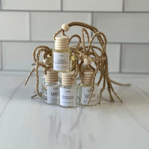 Può includere: Una collezione di piccole bottiglie di vetro trasparente con tappi di legno e cordoncini dorati, ciascuna etichettata con un nome di profumo diverso. Le bottiglie sono disposte insieme, suggerendo che siano per aromaterapia o profumo. Lo sfondo è piastrellato di bianco.