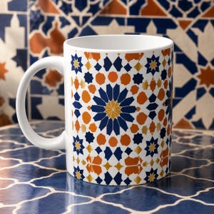 Op de afbeelding: Witte keramische mok met een geometrisch patroon in marineblauw, oranje en goud. Het ontwerp bevat ster- en ruitvormen. De mok heeft een wit handvat en staat tegen een achtergrond met patronen.