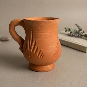 Könnte beinhalten: Ein Terrakotta-Becher mit matter Oberfläche, bestehend aus einem bauchigen Körper, einem stabilen Henkel und einem Standfuß. Die Außenseite ist mit geschnitzten, blattartigen vertikalen Mustern verziert, die gleichmäßig um den Umfang verteilt sind.