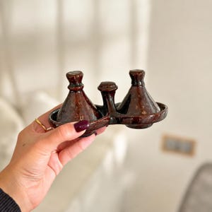 Può includere: Un set per condimenti in ceramica smaltata marrone scuro con due contenitori conici a forma di tajine collegati da un manico centrale. La finitura lucida riflette la luce e la base è integrata in un unico pezzo con un piccolo pomello.