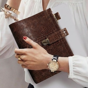 Puede incluir: Un diario de cuero marrón con un patrón de piel de serpiente, asegurado con un cierre. Se ve un reloj dorado con una correa de cuero negro y pulseras doradas. La persona lleva una blusa blanca.