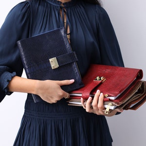 May include: A woman holds two leather-bound journals. One is navy blue with a snakeskin pattern and the other is red with a textured finish. Both have gold hardware. The red journal is stacked on top of several other journals.