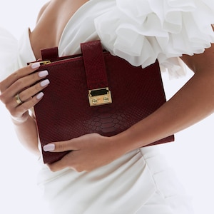 Puede incluir: Un clutch de piel de serpiente color burdeos con cierre de broche y cremallera dorados. El clutch lo lleva una persona que lleva un vestido blanco.