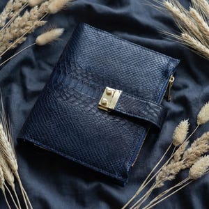 May include: A navy blue leather journal with a textured snakeskin pattern, a gold-colored clasp, and a zipper closure. The journal is surrounded by dried wheat stalks on a dark blue fabric background.