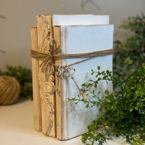 May include: A stack of three white books with decorative gold trim and a key tied with twine. The books are on a white surface with green foliage in the background.