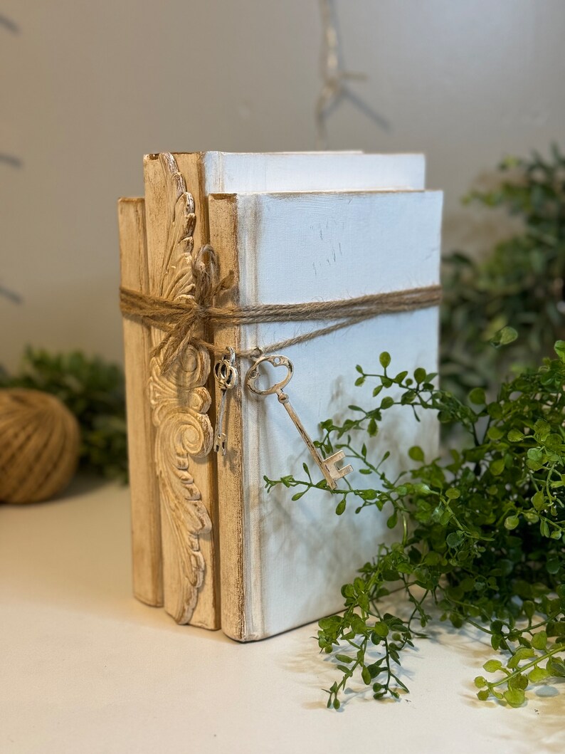 May include: A stack of three white books with decorative gold trim and a key tied with twine. The books are on a white surface with green foliage in the background.