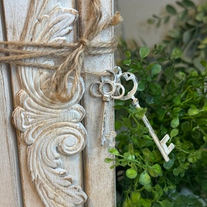May include: A white wooden frame with ornate details, tied with twine and decorated with antique keys. The frame is set against a backdrop of green foliage.