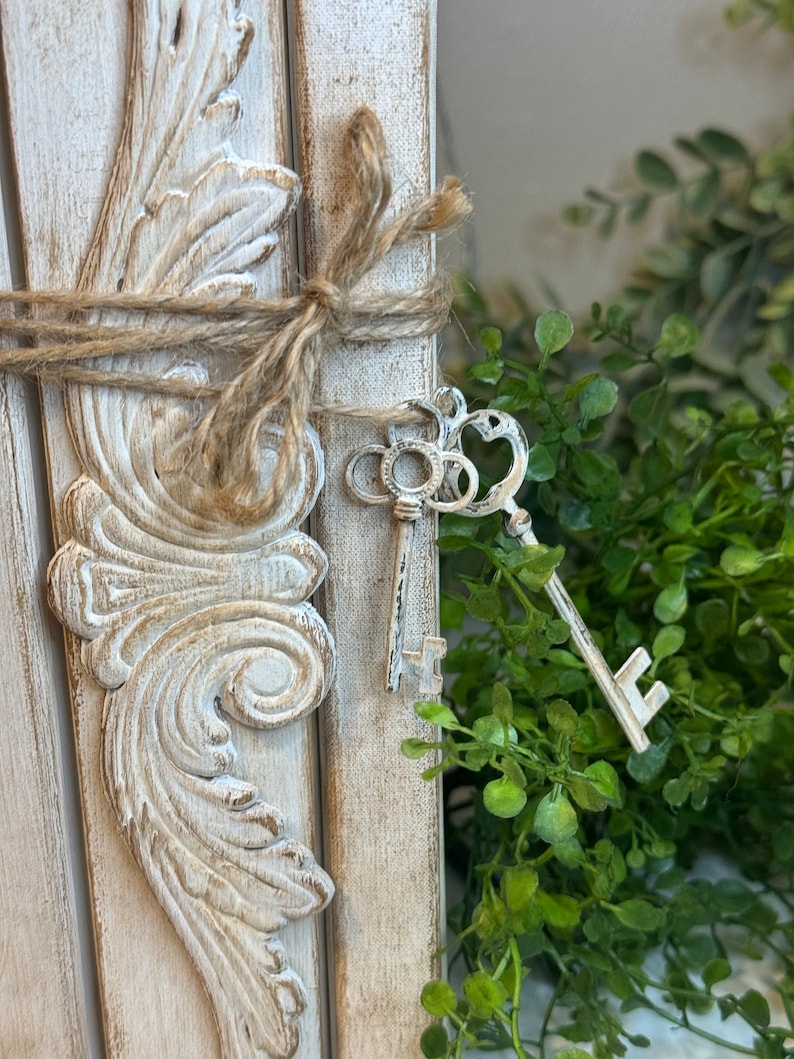 May include: A white wooden frame with ornate details, tied with twine and decorated with antique keys. The frame is set against a backdrop of green foliage.