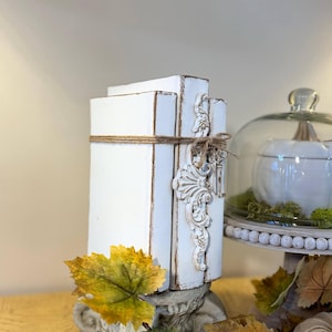 May include: Two white painted books stacked on top of each other with a decorative accent on the side. The books are tied together with twine and have a distressed look. The books are sitting on a decorative base with autumn leaves around them.