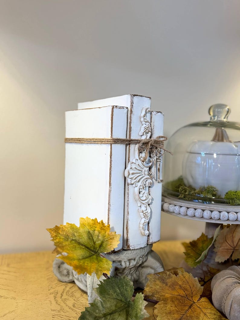 May include: Two white painted books stacked on top of each other with a decorative accent on the side. The books are tied together with twine and have a distressed look. The books are sitting on a decorative base with autumn leaves around them.