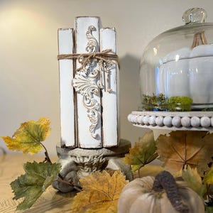 May include: A stack of white painted books with a decorative metal accent and twine tied around them. The books are sitting on a pedestal with a white pumpkin and autumn leaves in the foreground.