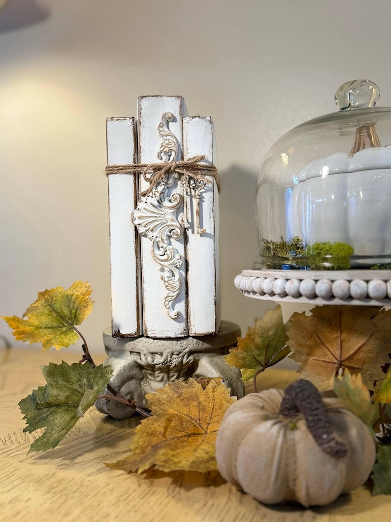 May include: A stack of white painted books with a decorative metal accent and twine tied around them. The books are sitting on a pedestal with a white pumpkin and autumn leaves in the foreground.