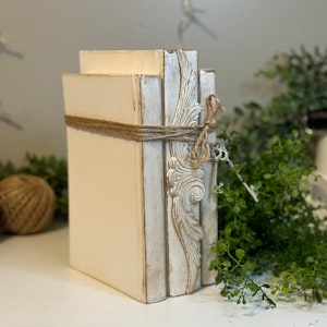 May include: A stack of three white decorative books with distressed paint and a decorative design on the spine. The books are tied together with twine.