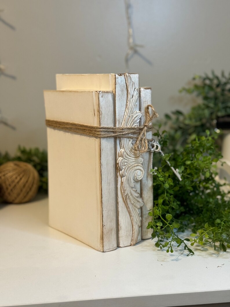 May include: A stack of three white decorative books with distressed paint and a decorative design on the spine. The books are tied together with twine.