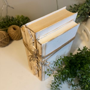May include: A stack of three white and gold decorative books with a key attached to the bottom book. The books are tied with twine and have a gold leaf design on the front.