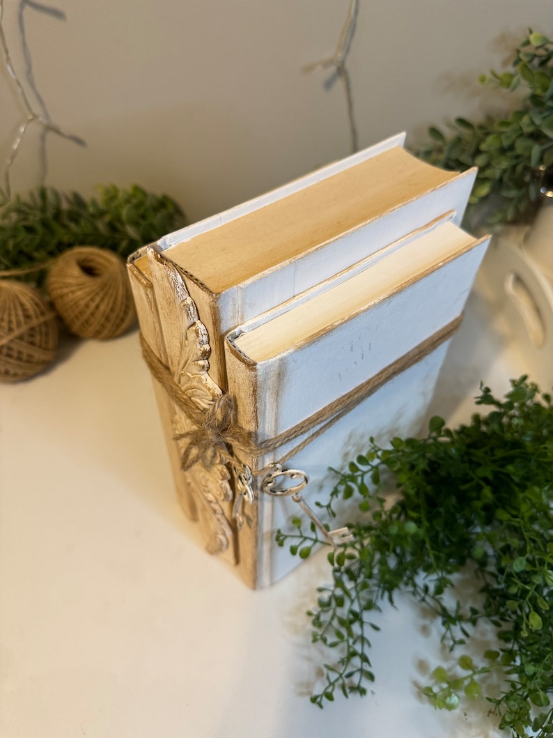 May include: A stack of three white and gold decorative books with a key attached to the bottom book. The books are tied with twine and have a gold leaf design on the front.
