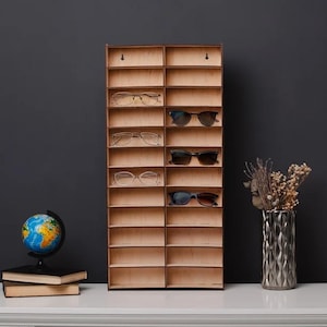 Puede incluir: Un expositor de gafas de madera con múltiples compartimentos, que contiene varios pares de gafas y gafas de sol. La pantalla está montada en una pared oscura, con un globo terráqueo, libros y un jarrón decorativo sobre una superficie blanca.