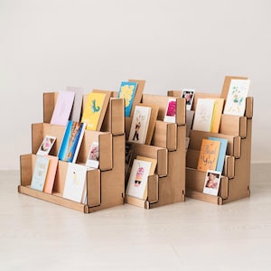 Könnte beinhalten: Drei Holz-Ausstellungsstände mit mehreren Ebenen, die jeweils verschiedene Grußkarten enthalten. Die Karten weisen farbenfrohe Designs und Texte auf. Die Ständer sind aus hellem Holz gefertigt und auf einer hellen Oberfläche angeordnet.