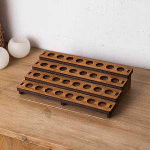 Op de afbeelding: Een houten displaystandaard met meerdere niveaus en talrijke ronde gaten. De standaard is gemaakt van gebeitst hout en heeft een warme bruine kleur. Het ontwerp heeft een trapsgewijze structuur, ideaal voor het organiseren en presenteren van kleine items. De standaard staat op een houten oppervlak.