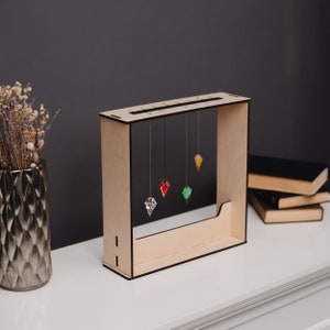 Puede incluir: Una pieza de arte cinético de madera con un marco rectangular. Cinco colgantes coloridos en forma de gema cuelgan de la parte superior. Los colgantes son transparentes, rojos, verdes y amarillos. La pieza está sobre una superficie blanca.