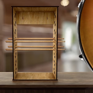 Pode incluir: Um suporte de madeira para baquetas, de forma retangular, com vários pinos horizontais para guardar as baquetas. O suporte é feito de madeira clara com uma borda mais escura. O fundo está desfocado, com um tambor visível à direita.