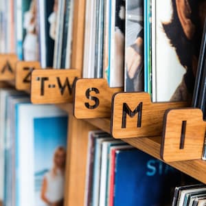 May include: A set of wooden alphabet dividers for vinyl record storage, featuring laser-cut letters including T-W, S, M, and I. These rectangular wooden tabs are designed to organize collections on shelves, with each letter clearly engraved.