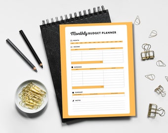 May include: A yellow monthly budget planner with sections for income, expenses, summary, and notes. The planner is on a black notebook with a spiral binding.