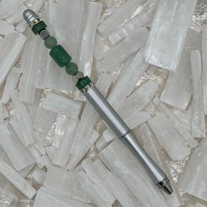 May include: A silver pen with a decorative top featuring green beads and accents. The pen is set against a background of white, crystalline formations, creating a contrast of textures and colours. The pen's design suggests a focus on writing and stationery.