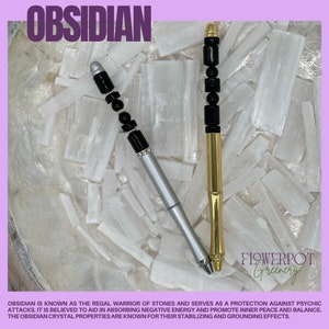 May include: Two obsidian crystal pens with black beads and silver and gold accents. The pens are resting on a bed of white selenite crystals. The text "OBSIDIAN" is at the top of the image and "FLOWERPOT Greenery" is at the bottom of the image.