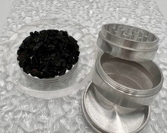 May include: A silver metal grinder with a black plastic lid and a black plastic tray. The grinder has a mesh screen and a compartment for collecting kief. The tray is filled with black, irregularly shaped pieces of herb.