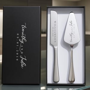 May include: A silver cake knife and server set in a black gift box. The knife and server are engraved with names and a date. The box is labeled with names and a date in a script font.
