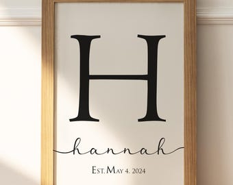 May include: A framed print with a black letter H and the name "hannah" written in cursive below. The text "Est. May 4, 2024" is printed below the name.