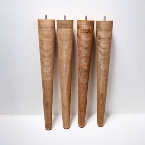 May include: Four light brown wooden furniture legs with a tapered, cylindrical shape and a fine, horizontal ribbed texture. Each leg features a small, silver-coloured metal threaded bolt protruding from the top for attachment to furniture.