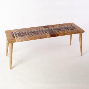 Puede incluir: Banco de madera con asiento tejido. El banco tiene cuatro patas cónicas y un asiento rectangular. El asiento está hecho de tiras tejidas de materiales marrón claro y oscuro. El marco es de madera de color claro.