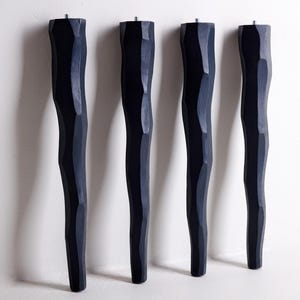 May include: A set of four black furniture legs with a faceted, hand-carved appearance. Each leg features a vertical, irregular, and textured surface with a metallic threaded bolt protruding from the flat top for attachment to furniture bases.