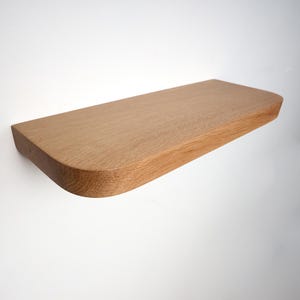 Puede incluir: Una estantería flotante de madera marrón claro montada en una pared blanca. La estantería tiene forma rectangular con esquinas redondeadas. La veta de la madera es visible y parece ser de madera maciza.