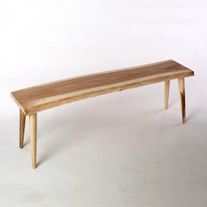 May include: A light brown wooden bench with four tapered legs. The bench has a rectangular top with a lighter wood border and a darker wood center. The legs are angled outwards, providing stability.