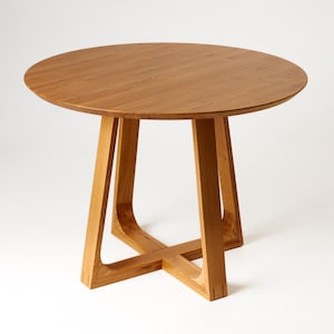 Puede incluir: Una mesa redonda de madera con acabado marrón claro. La mesa tiene una tapa circular y un diseño de base único con cuatro patas anguladas que se cruzan en la parte inferior. La mesa tiene aproximadamente 76 cm de diámetro.