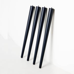 May include: Four tapered, black wooden furniture legs lean against a white wall. Each leg has a slightly rounded top and a narrow, pointed base. The legs are evenly spaced and appear to be new.